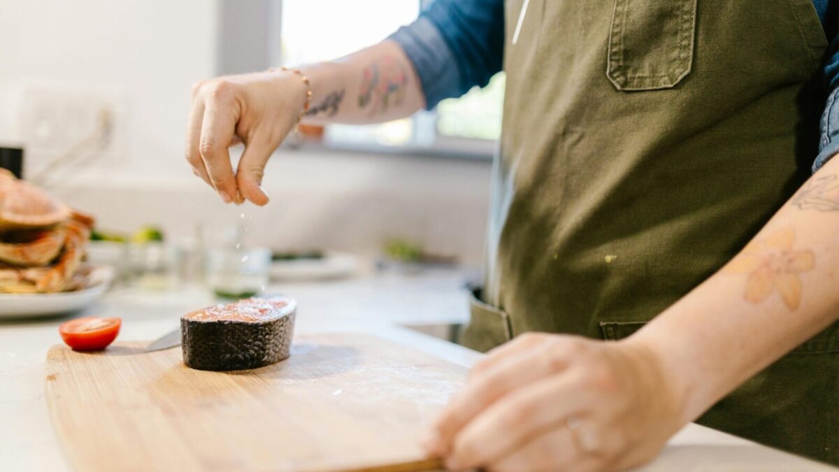 Close-up of a tattooed chef seasoning a salmon steak with salt in a modern kitchen.