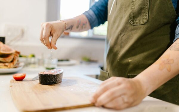 Close-up of a tattooed chef seasoning a salmon steak with salt in a modern kitchen.