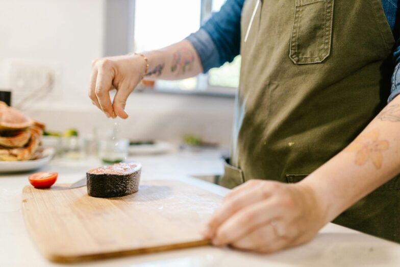 Close-up of a tattooed chef seasoning a salmon steak with salt in a modern kitchen.
