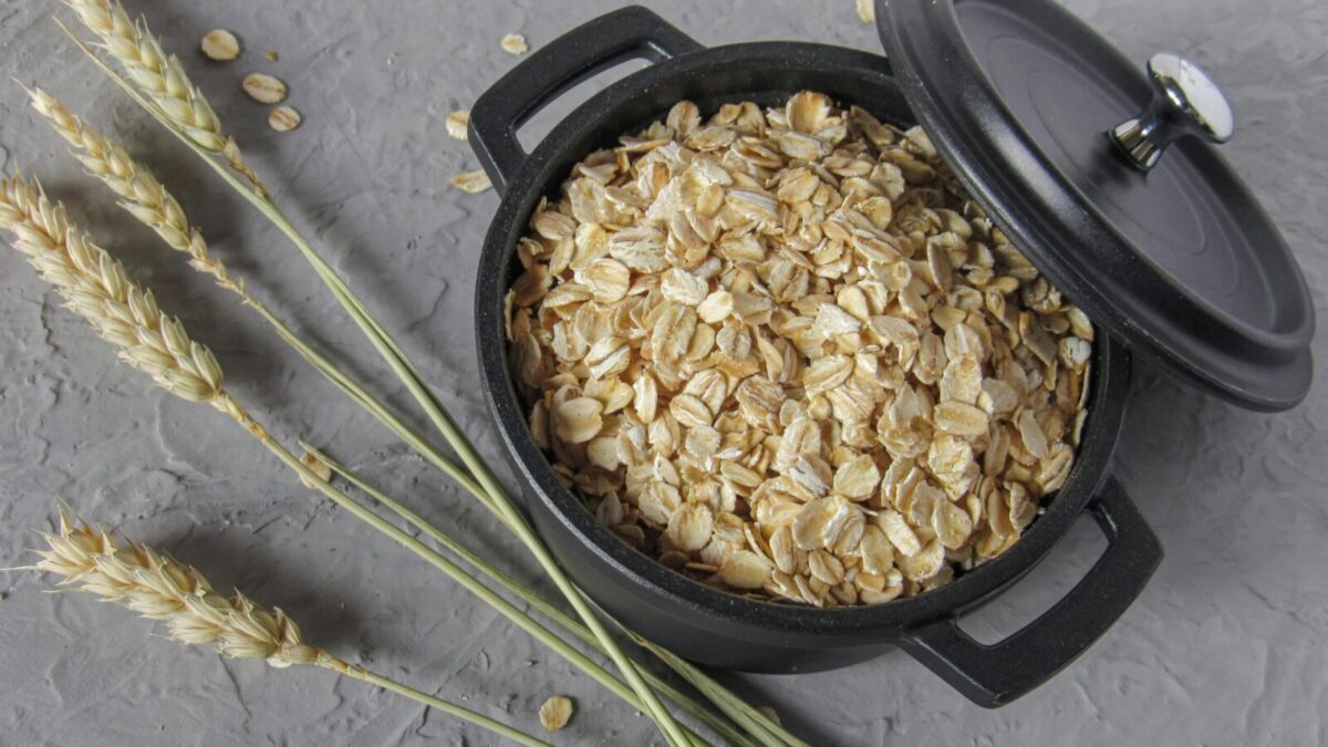 Flat lay of rolled oats in a black pot surrounded by wheat stalks on a textured surface.