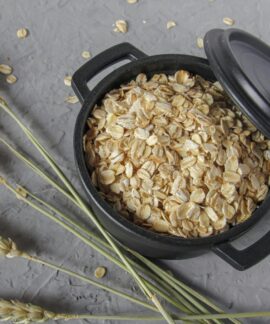 Flat lay of rolled oats in a black pot surrounded by wheat stalks on a textured surface.
