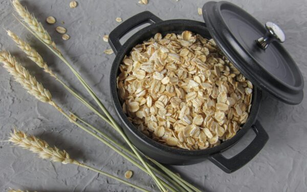 Flat lay of rolled oats in a black pot surrounded by wheat stalks on a textured surface.