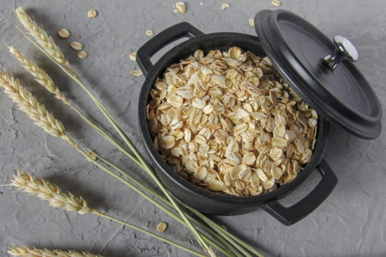 Flat lay of rolled oats in a black pot surrounded by wheat stalks on a textured surface.