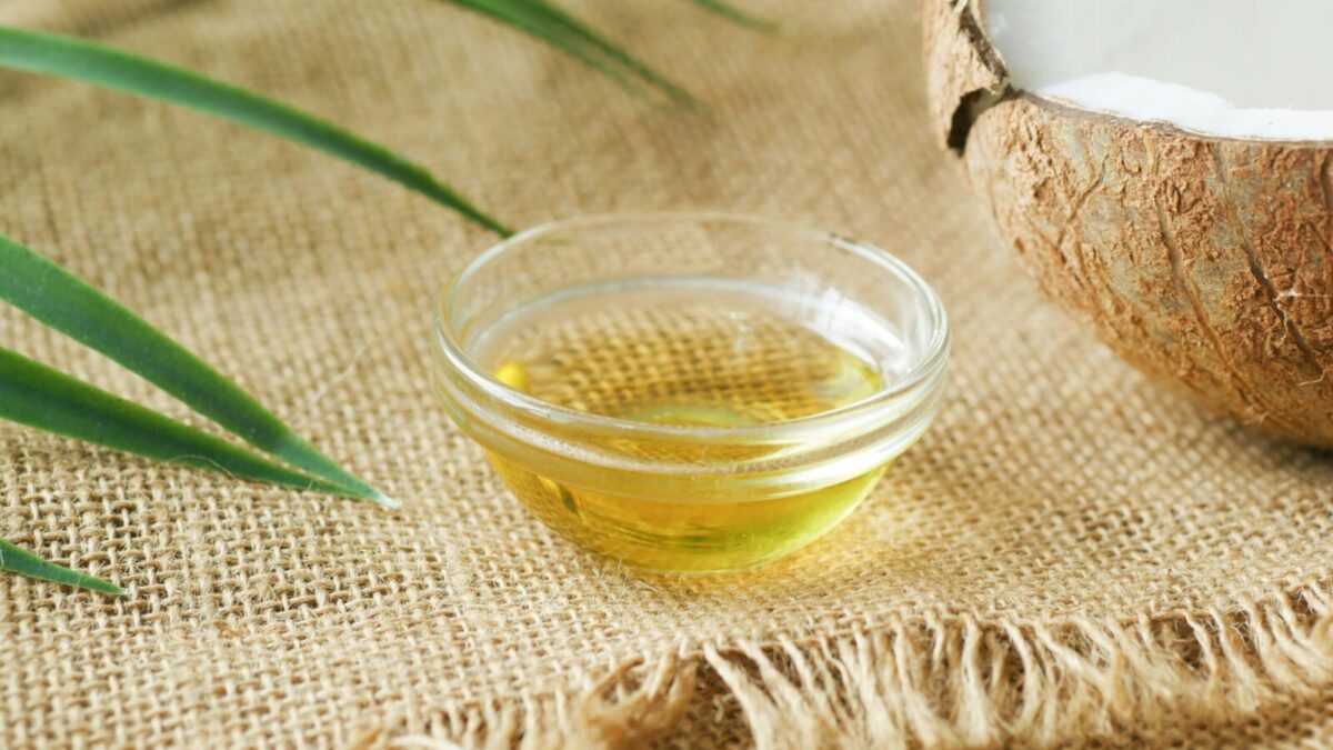 A close-up of coconut oil in a glass bowl on burlap, surrounded by coconut shell and leaves.
