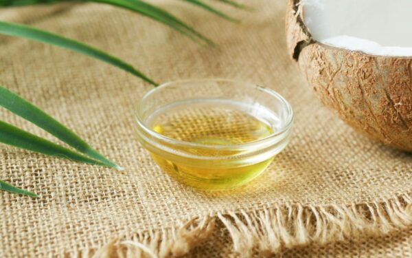 A close-up of coconut oil in a glass bowl on burlap, surrounded by coconut shell and leaves.