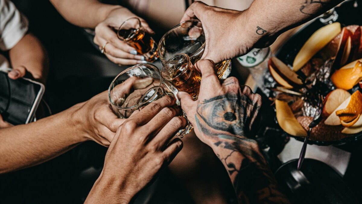 A close-up of friends clinking glasses in a festive setting, highlighting tattoos and diverse hands.