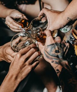A close-up of friends clinking glasses in a festive setting, highlighting tattoos and diverse hands.