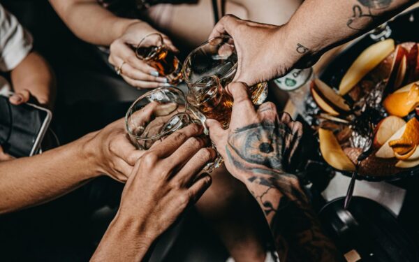 A close-up of friends clinking glasses in a festive setting, highlighting tattoos and diverse hands.