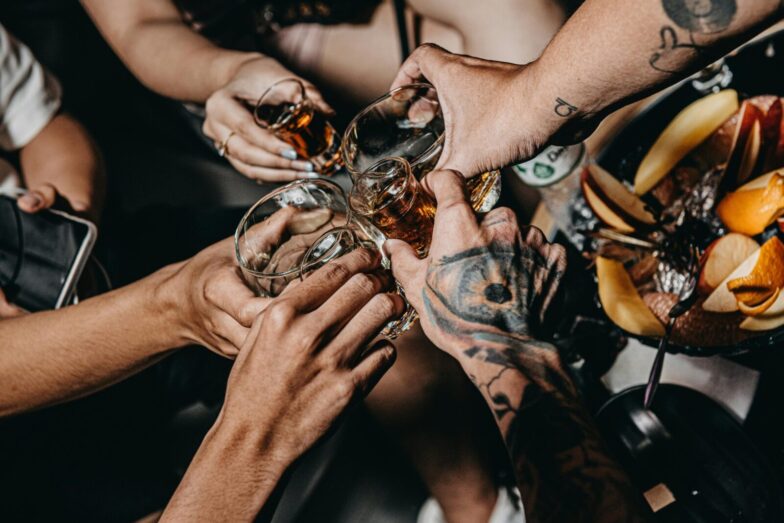 A close-up of friends clinking glasses in a festive setting, highlighting tattoos and diverse hands.