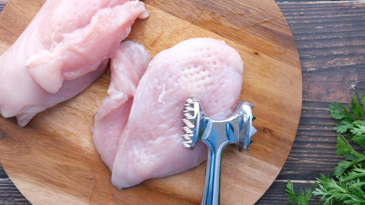 Top view of raw chicken breasts on a wooden board with a meat mallet for tenderizing.