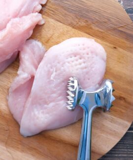 Top view of raw chicken breasts on a wooden board with a meat mallet for tenderizing.