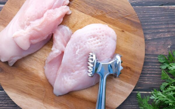 Top view of raw chicken breasts on a wooden board with a meat mallet for tenderizing.