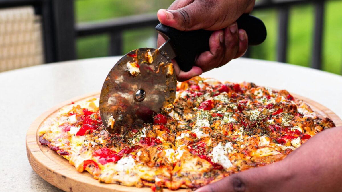 Close-up of hands slicing fresh pizza with cutter outdoors, vibrant toppings.