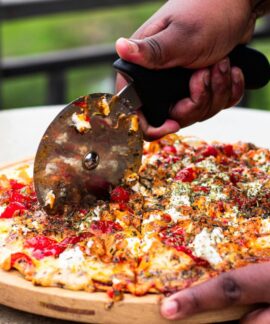 Close-up of hands slicing fresh pizza with cutter outdoors, vibrant toppings.