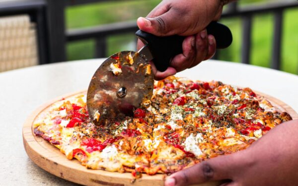 Close-up of hands slicing fresh pizza with cutter outdoors, vibrant toppings.