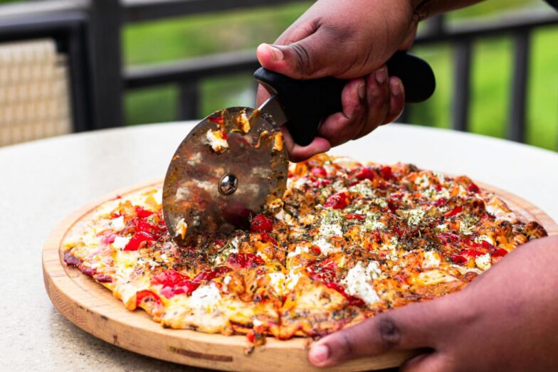 Close-up of hands slicing fresh pizza with cutter outdoors, vibrant toppings.
