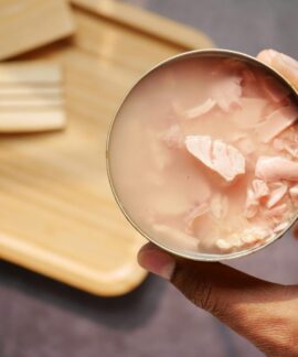 A close-up of an opened canned tuna held over a wooden cutting board and utensils.