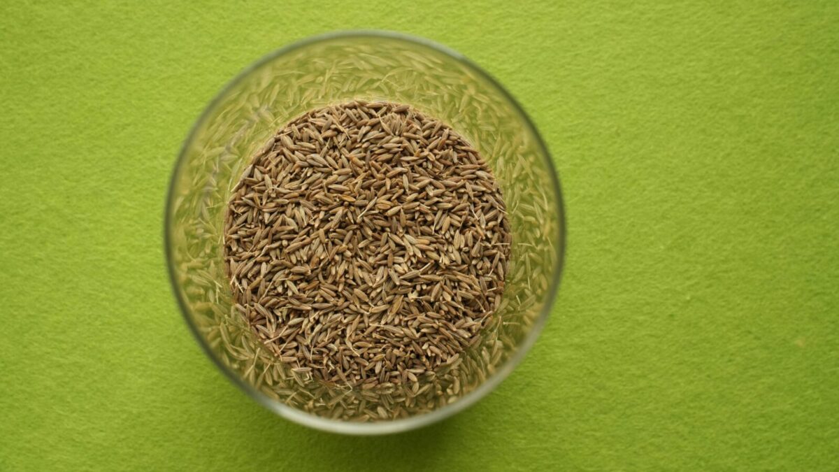 Cumin seeds in a clear glass bowl on a vibrant green background, viewed from above.