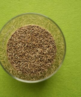 Cumin seeds in a clear glass bowl on a vibrant green background, viewed from above.