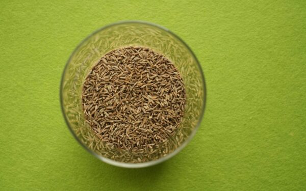 Cumin seeds in a clear glass bowl on a vibrant green background, viewed from above.