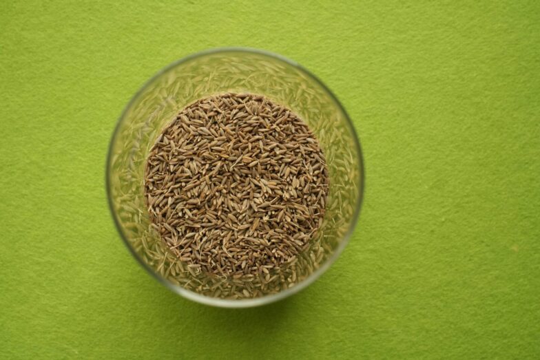 Cumin seeds in a clear glass bowl on a vibrant green background, viewed from above.