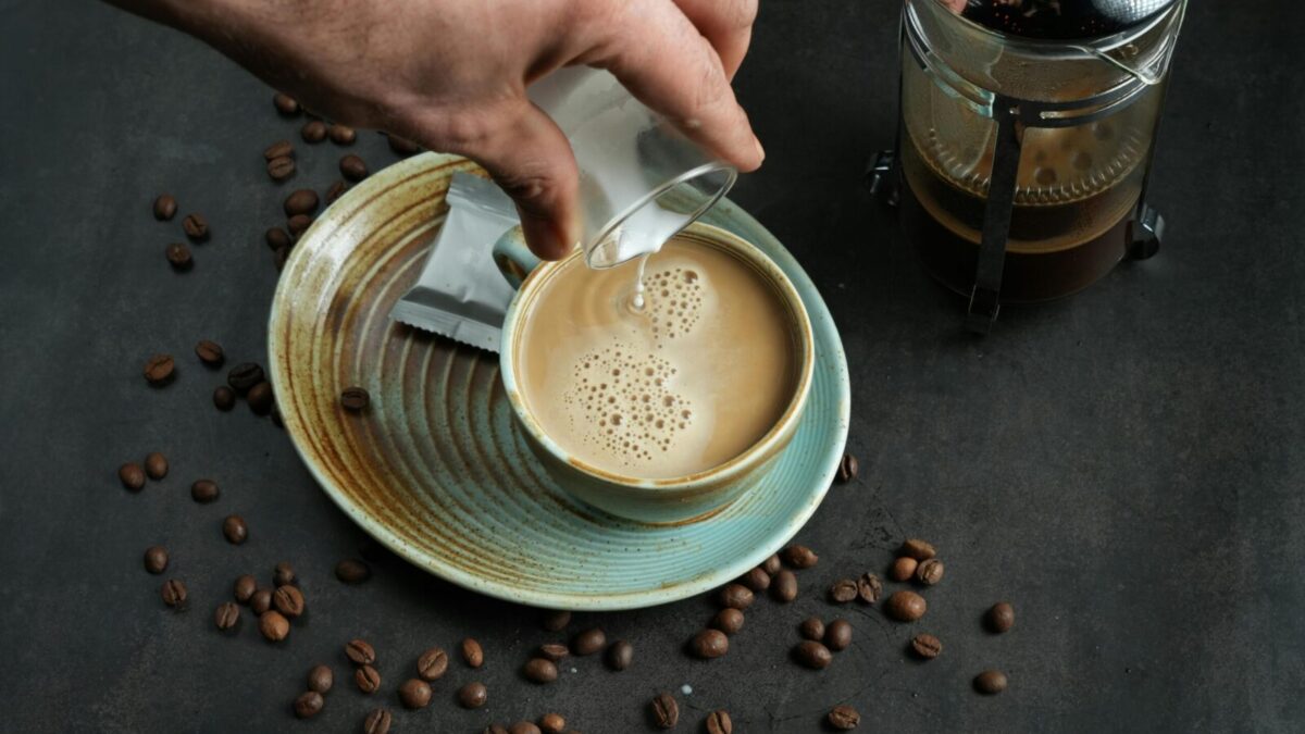 Close-up of coffee with milk pouring and French press on a dark surface.