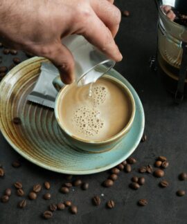 Close-up of coffee with milk pouring and French press on a dark surface.