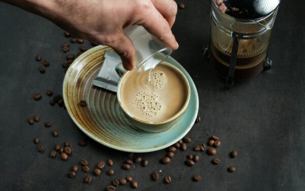 Close-up of coffee with milk pouring and French press on a dark surface.