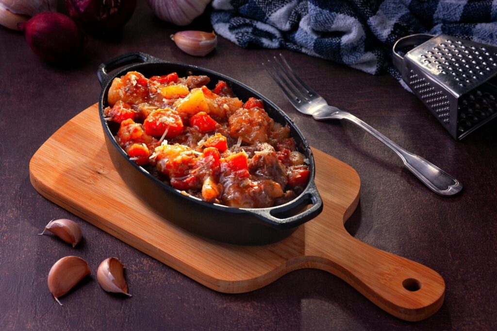 Savory vegetable stew served in cast iron on wooden board, paired with garlic cloves and kitchen utensils.