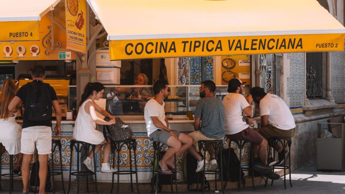 People enjoying food at a Valencian street stall under a bright yellow awning.