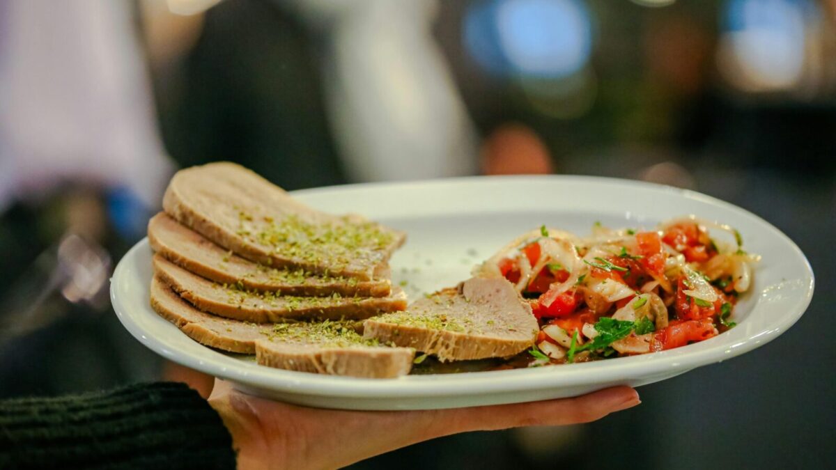 Close-up of a gourmet dish featuring beef tongue slices and fresh tomato salsa held by a server indoors.