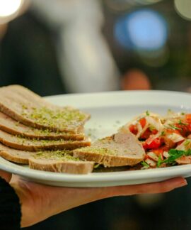 Close-up of a gourmet dish featuring beef tongue slices and fresh tomato salsa held by a server indoors.