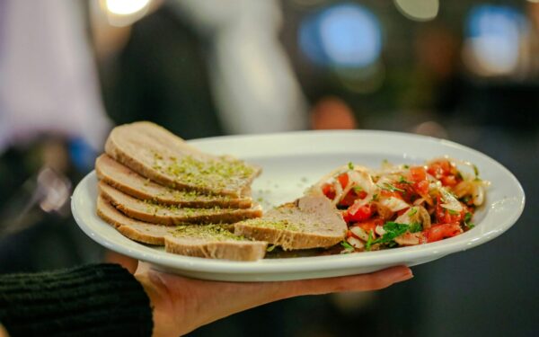 Close-up of a gourmet dish featuring beef tongue slices and fresh tomato salsa held by a server indoors.