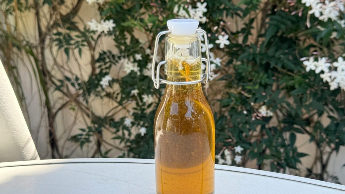 A glass bottle of olive oil placed on a table outdoors with white flowers in the background.