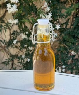 A glass bottle of olive oil placed on a table outdoors with white flowers in the background.
