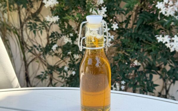 A glass bottle of olive oil placed on a table outdoors with white flowers in the background.