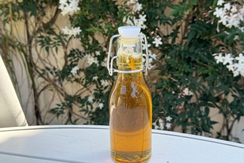 A glass bottle of olive oil placed on a table outdoors with white flowers in the background.