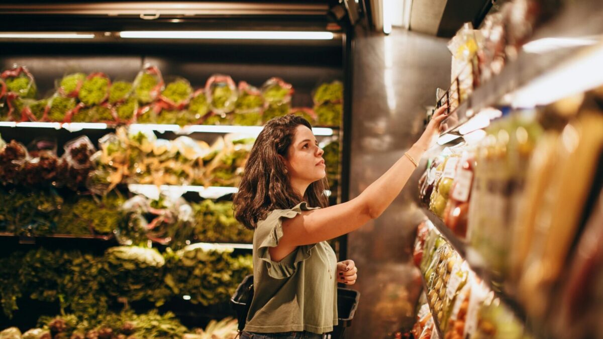 A woman selects fresh vegetables in a well-lit supermarket produce aisle.