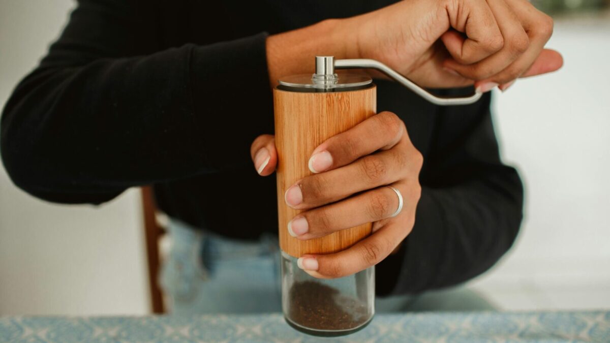 A person grinding coffee beans using a manual wooden coffee grinder on a patterned tablecloth.