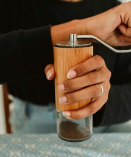 A person grinding coffee beans using a manual wooden coffee grinder on a patterned tablecloth.