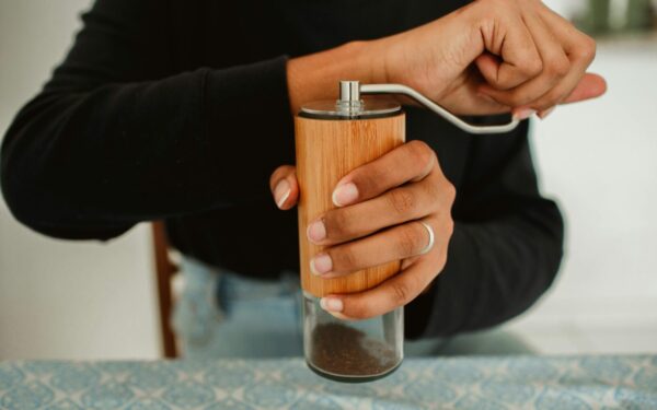 A person grinding coffee beans using a manual wooden coffee grinder on a patterned tablecloth.
