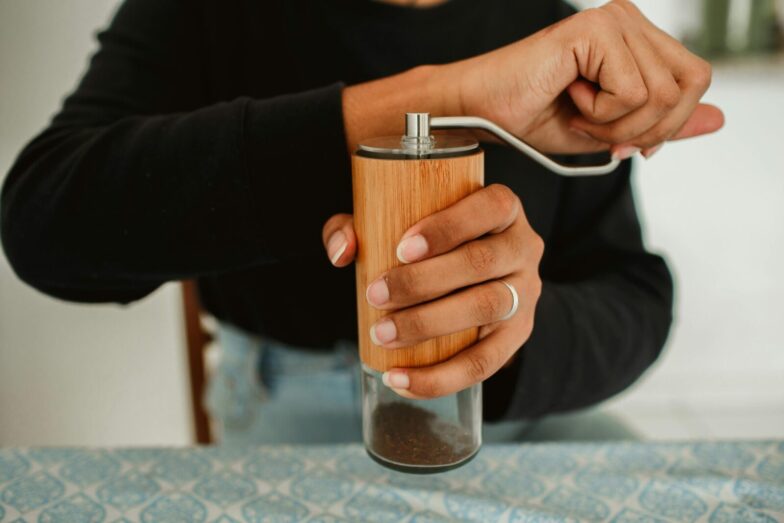 A person grinding coffee beans using a manual wooden coffee grinder on a patterned tablecloth.
