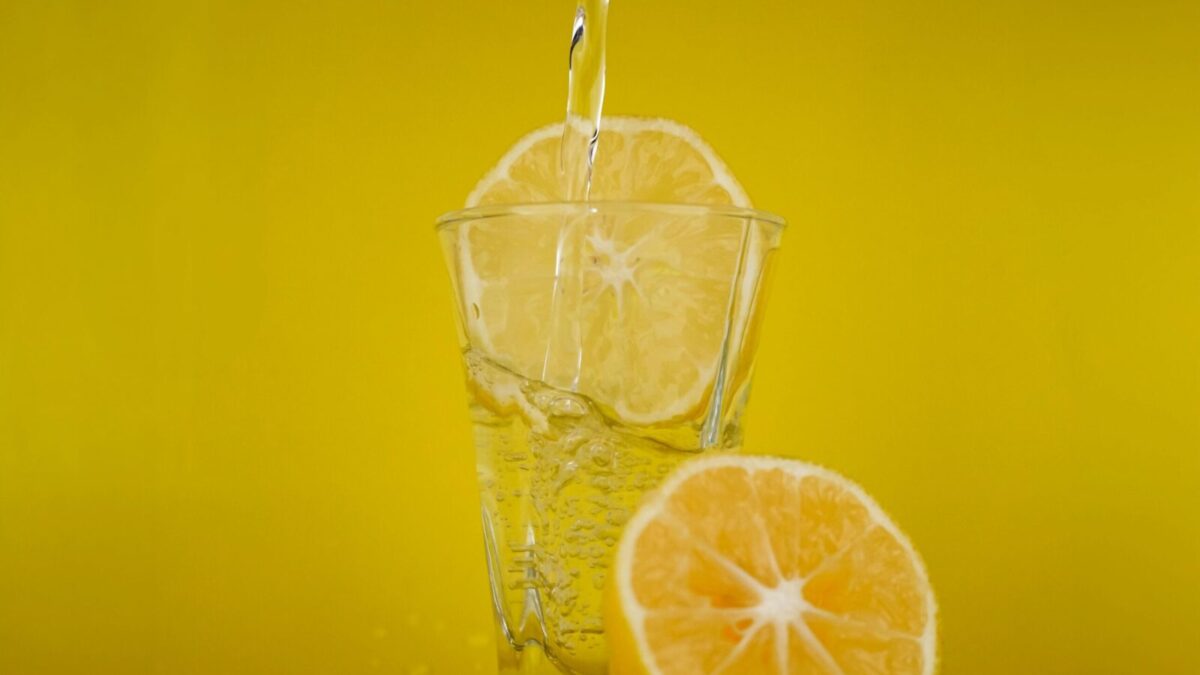 A close-up of water pouring into a glass with lemon slices, vibrant yellow backdrop.