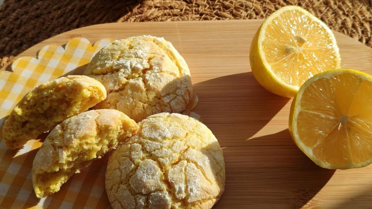 Freshly baked lemon cookies with halved lemons on a wooden platter in sunlight.
