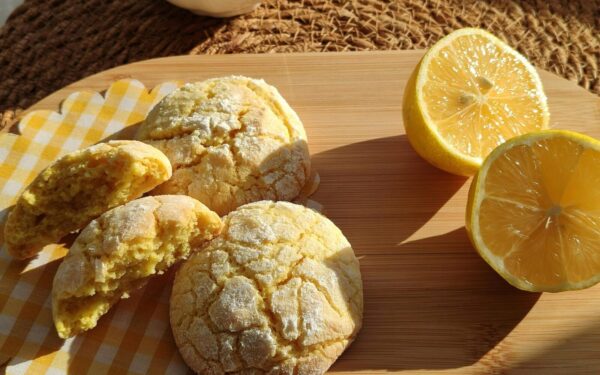 Freshly baked lemon cookies with halved lemons on a wooden platter in sunlight.