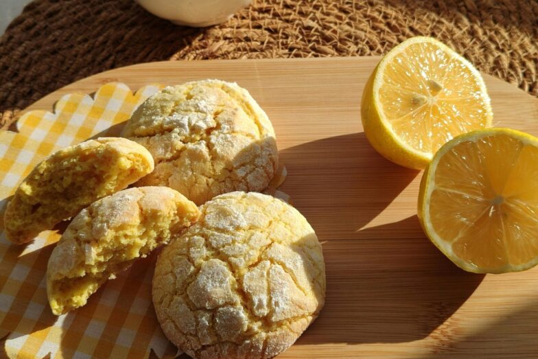 Freshly baked lemon cookies with halved lemons on a wooden platter in sunlight.