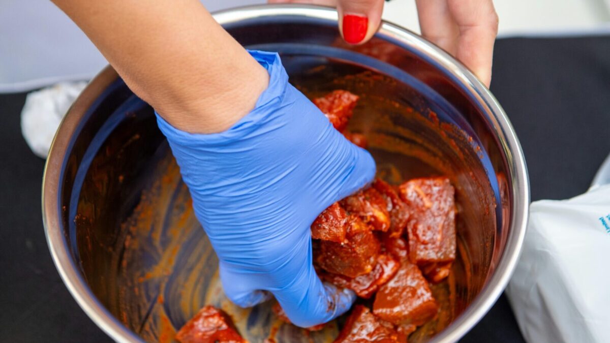 Close-up of marinated meat being prepared with blue gloves in a stainless steel bowl.