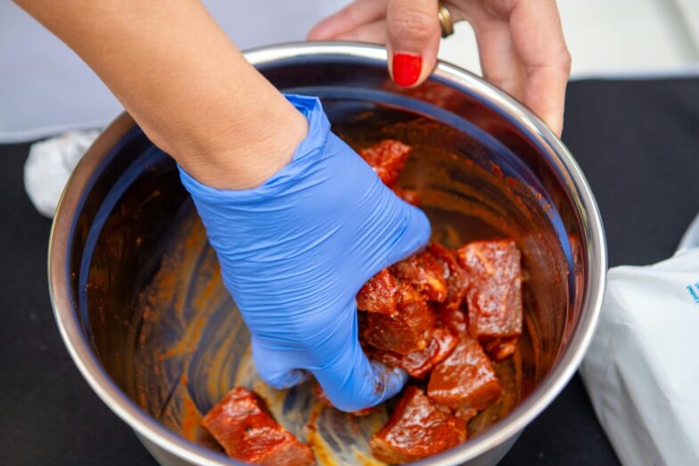Close-up of marinated meat being prepared with blue gloves in a stainless steel bowl.