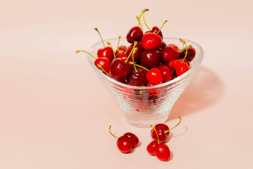 A vibrant display of fresh red cherries in a glass bowl against a soft pink background.