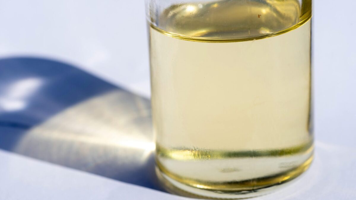 A close-up image of a glass bottle with liquid, showcasing light reflection and shadow on a white surface.